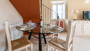 A dining area with a glass table, wicker chairs, and a view of a kitchen with a TV and books.