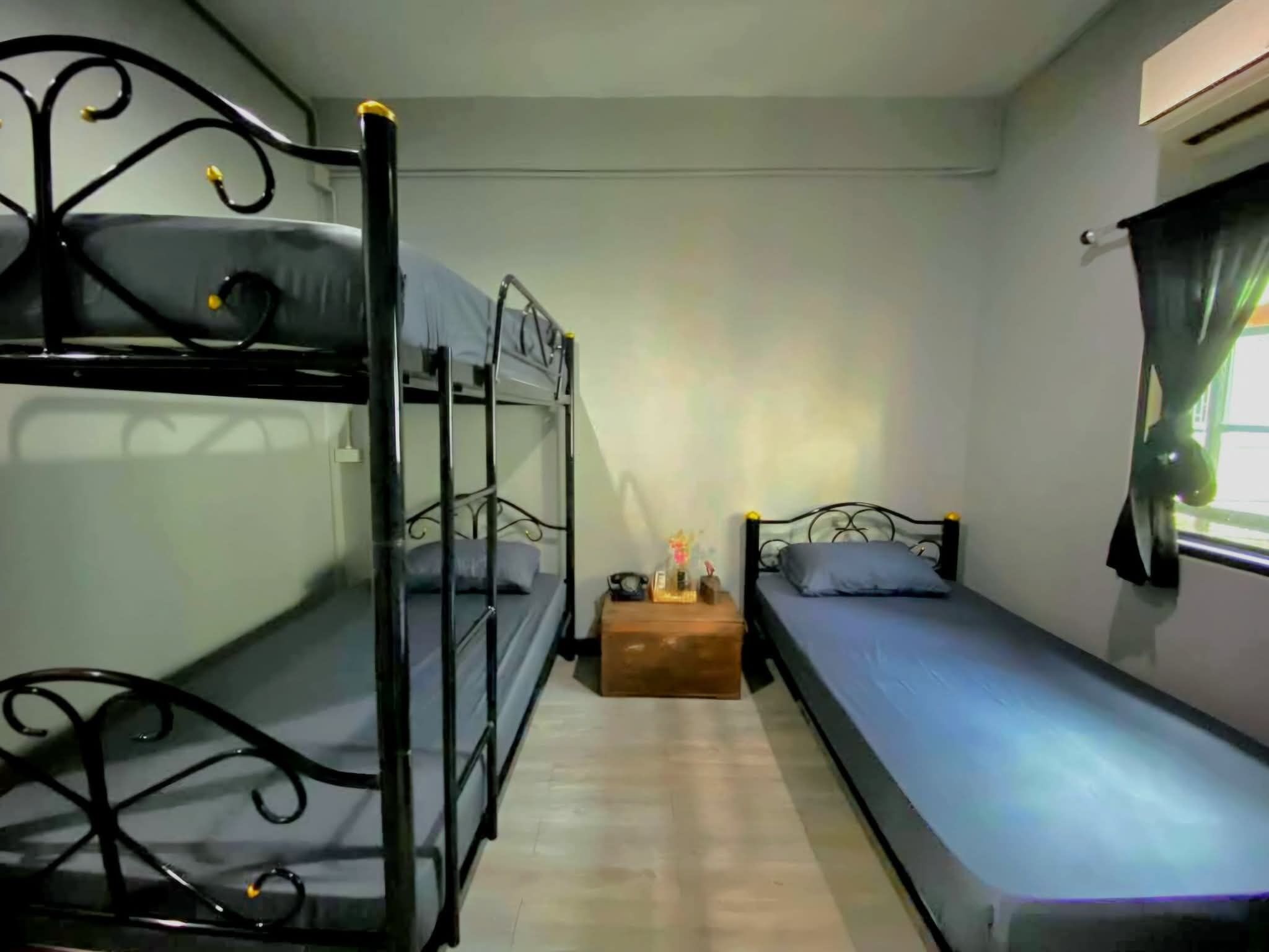 A dormitory room with bunk beds and a single bed, a small wooden nightstand, and a window with curtains.