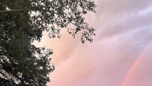 A large, dark-colored house with a prominent triangular roof and a balcony, surrounded by a grassy field and trees, under a cloudy sky with a visible rainbow. - Charming 3BR/2BA Retreat | Pond | Fire Pit | Pets (Skaneateles)