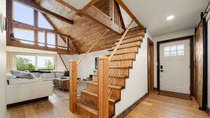 A modern interior with a wooden staircase, a living area with a sofa, and a view through large windows. - Charming 3BR/2BA Retreat | Pond | Fire Pit | Pets (Skaneateles)