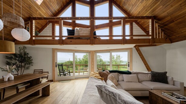 A spacious loft-style room with a high ceiling, wooden beams, a large sliding glass door leading to an outdoor deck, and a cozy seating area with a sofa and a bench. - Charming 3BR/2BA Retreat | Pond | Fire Pit | Pets (Skaneateles)