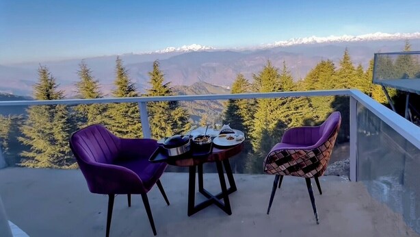 Ein Balkon mit zwei violetten Stühlen und einem kleinen Tisch, mit Blick auf eine gebirgige Landschaft mit schneebedeckten Gipfeln und dichtem Wald.
