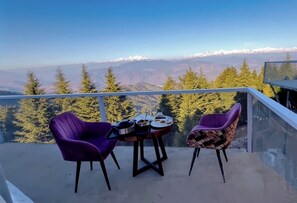 A balcony with two purple chairs and a small table, overlooking a mountainous landscape with snow-capped peaks and dense forest. - Nilayam Resorts (Tehri)
