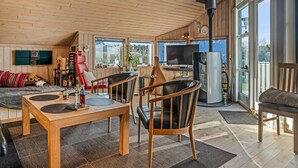 Amplia sala de estar con techo de madera, una mesa de comedor y varias sillas. Hay un televisor, una chimenea y ventanas grandes que ofrecen luz natural.