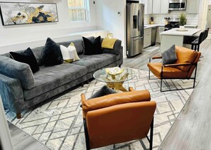A modern living room with a grey sofa, a glass coffee table, and two leather armchairs.