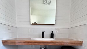 Ein Badezimmer mit weiß gefliesten Wänden, einem hölzernen Waschtisch mit Waschbecken und einem Spiegel darüber. Es gibt zwei Handtuchhalter mit gefalteten Handtüchern.