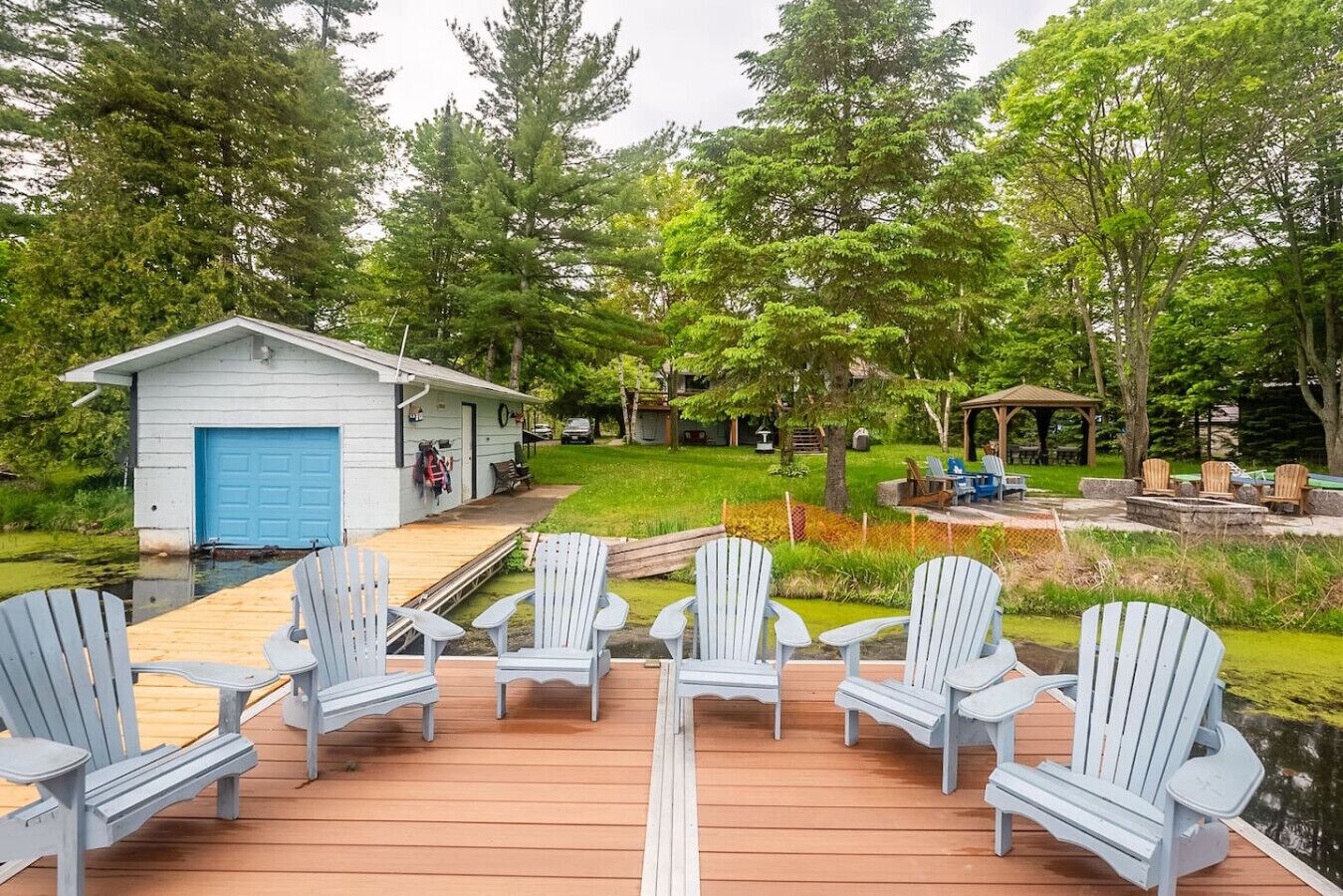 A deck with Adirondack chairs, a small shed with a blue door, and a gazebo in a wooded backyard.