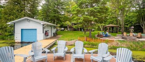 A deck with Adirondack chairs, a small shed with a blue door, and a gazebo in a wooded backyard.