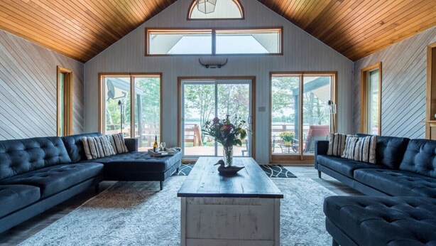 Ein geräumiges Wohnzimmer mit hoher Decke, großen Fenstern und zwei schwarzen Sofas.