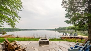 Eine Terrasse am Seeufer mit Adirondack-Stühlen, einer Feuerstelle und einem Steg mit Booten.
