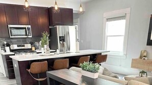 A modern kitchen with dark wood cabinets, a stainless steel refrigerator, a microwave, and a built-in oven. There is a central island with stools, a large window, and a seating area with a sofa and a coffee table. - Luxe Victorian 2BR/2BA Downtown w/Serene Backyard (Sacramento)