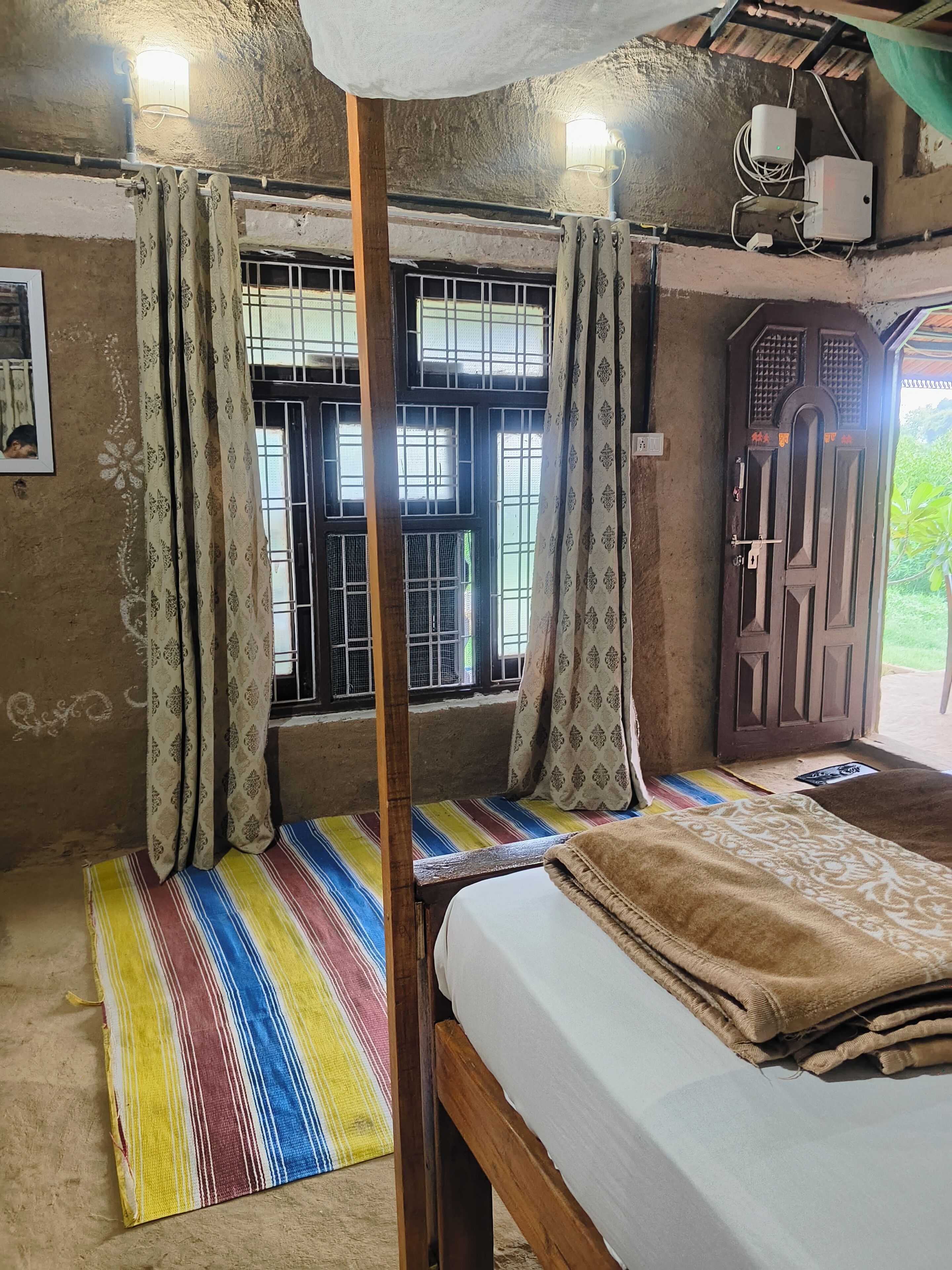Una habitación con una cama, una ventana con barrotes, una cortina, una lámpara en la pared y una puerta.