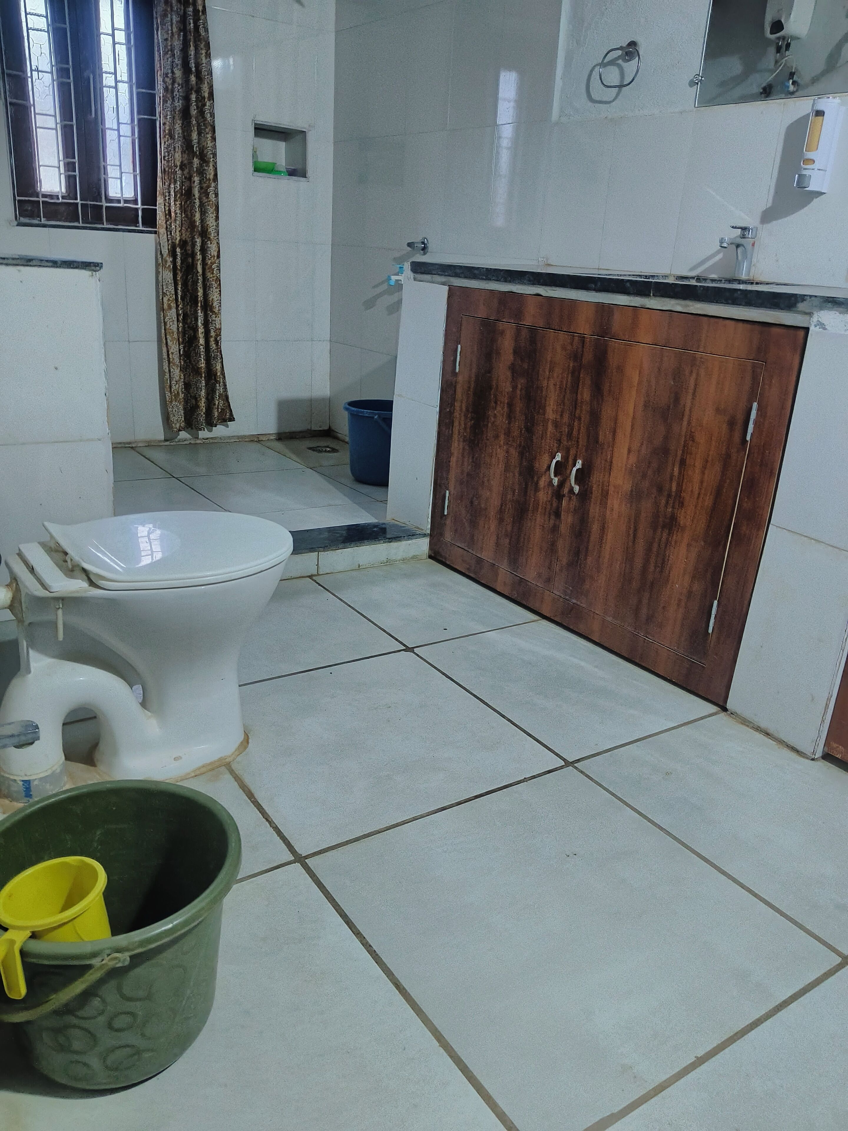 Un baño con inodoro, un armario de madera, un balde y una ventana con barrotes.