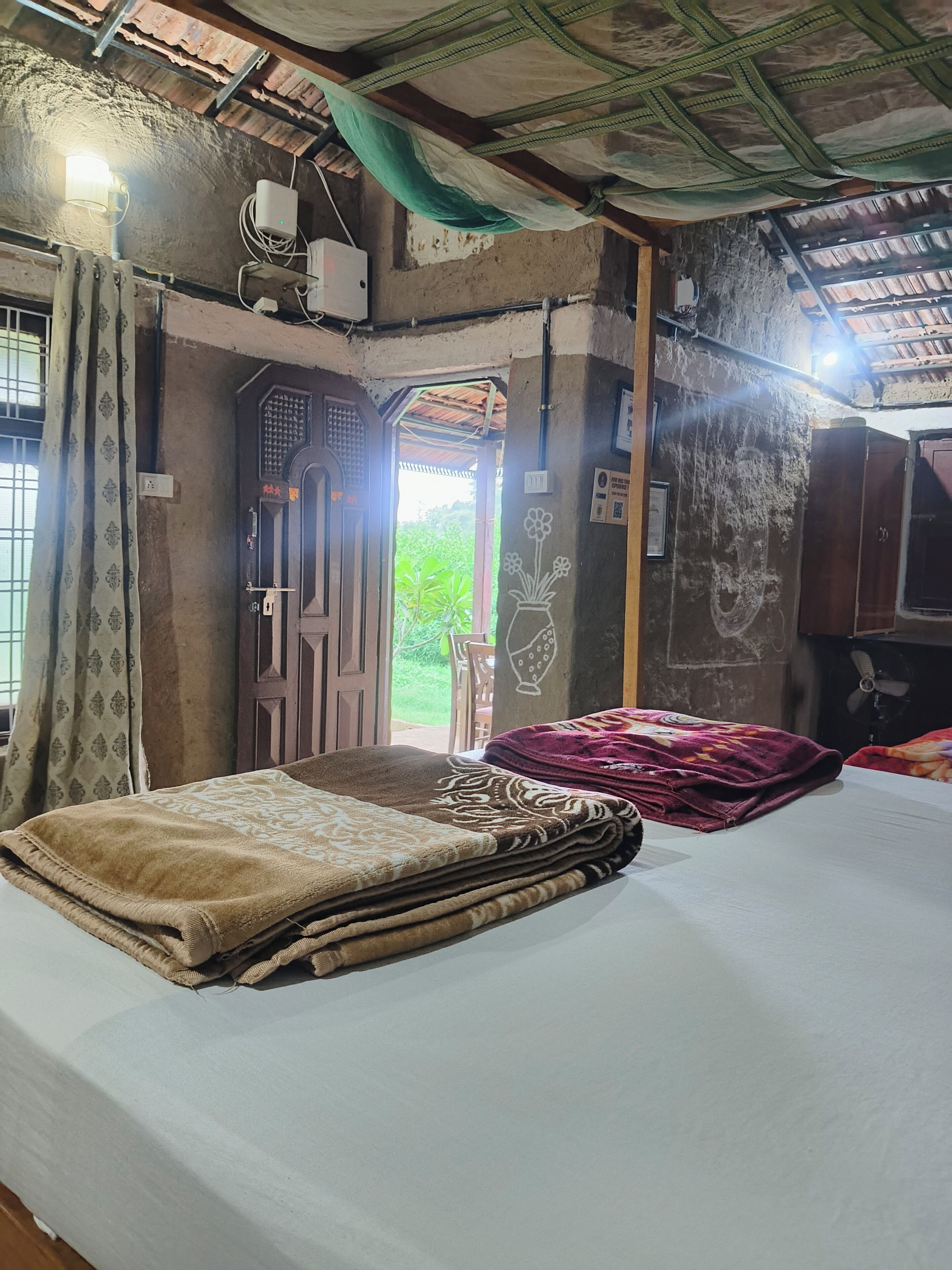 Ein Zimmer mit einem Bett, einer zur Außenwelt führenden Tür, einem Ventilator und einer Wand mit einer Blumendarstellung.