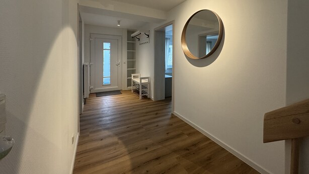 Een gang met houten vloer, een witte deur en een ronde spiegel aan de muur.
