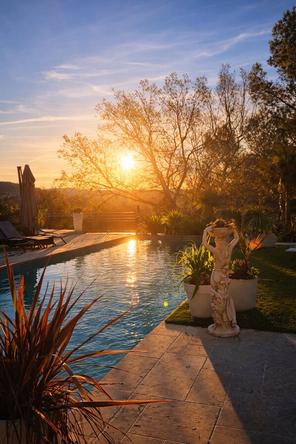 A swimming pool with a statue, surrounded by plants and trees, during sunset.