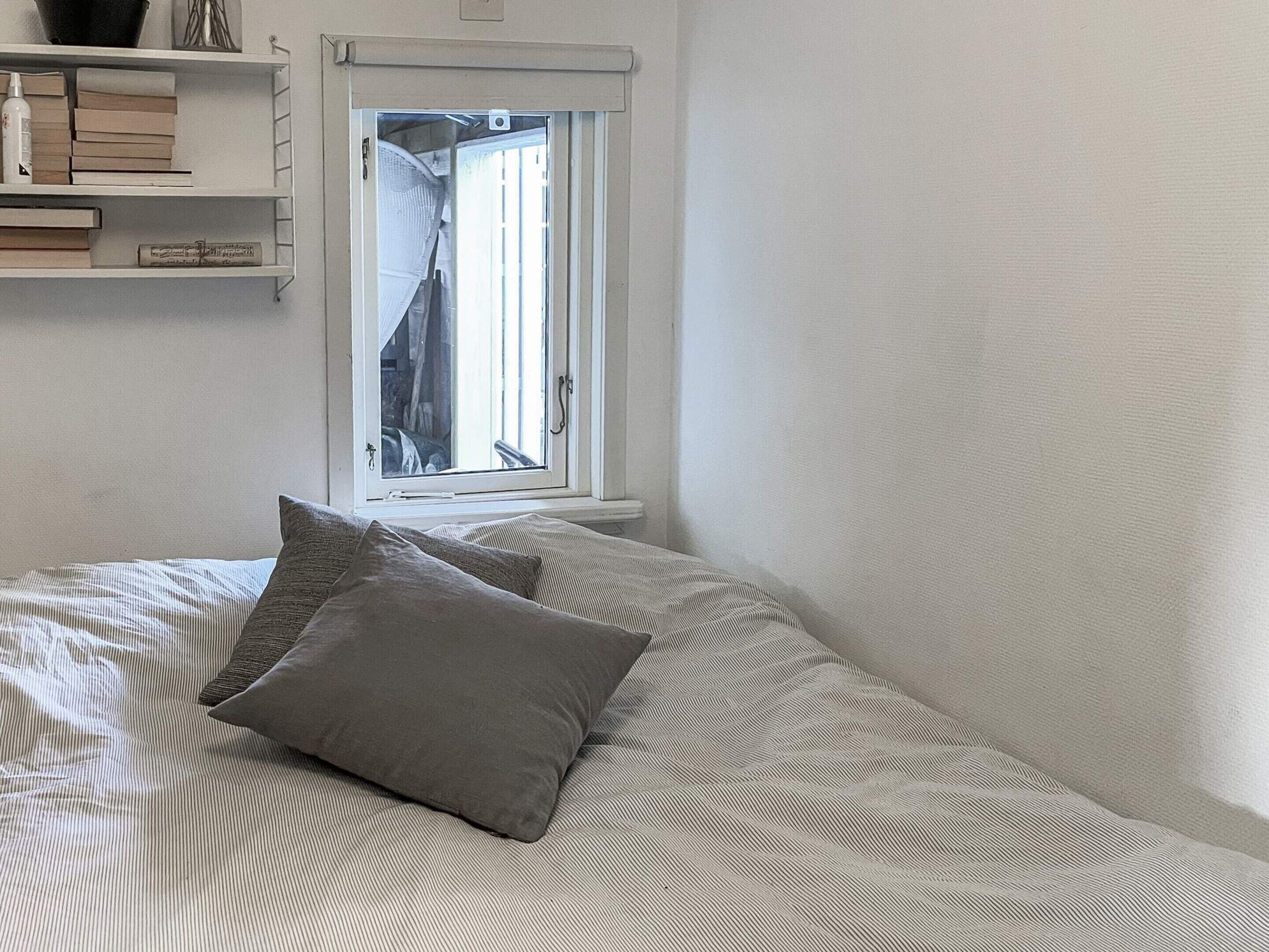 A bedroom with a bed, pillows, a window, and a shelf with books and a bottle.