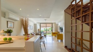 A modern kitchen with a central island, a dining area with yellow chairs, and a staircase with a wooden railing.