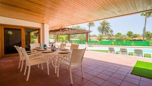 Un patio techado con techo de madera, zona de comedor al aire libre con mobiliario de ratán blanco, y vistas a una piscina y zonas verdes.