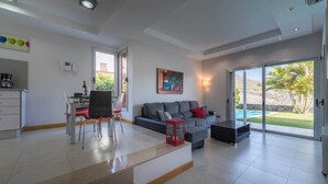 Un salon moderne avec un canapé gris, un coin repas comprenant une table et des chaises, et une grande porte-fenêtre coulissante donnant sur une zone avec piscine.