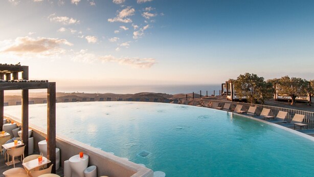 Une piscine à débordement offrant une vue sur la mer et les collines lointaines, entourée de chaises longues et de tables.
