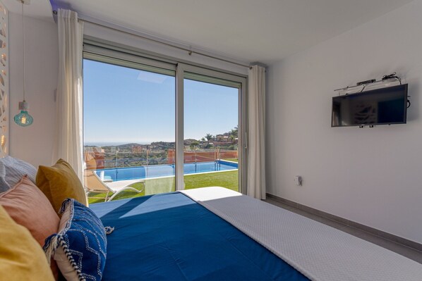 A bedroom with a large window, a bed with blue and white bedding, a TV on the wall, and a view of a pool and landscape.
