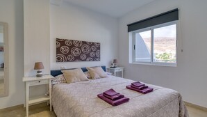 A bedroom with a bed, two nightstands, a lamp, a window with a view, and a decorative wall piece. - Apartment in Mogán Playa near the Beach (Mogán)