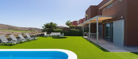 Un área de piscina moderna con césped artificial, sillas de descanso y un edificio con ventanas amplias y un porche cubierto.