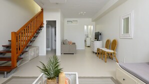 Ein geräumiges Wohnzimmer mit einer Holztreppe, einem grauen Sofa, Korbstühlen und einem kleinen Tisch mit einer Pflanze.