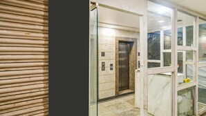 Ein Aufzug mit Marmorwand und Glastür, neben einer geschlossenen Aufzugtür mit Metallgriff.