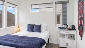 Une chambre avec un lit, deux fenêtres équipées de stores, une petite armoire blanche avec une bouilloire, et une affiche accrochée au mur.