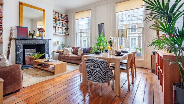 A spacious living room with a fireplace, wooden flooring, a dining area with a table and chairs, and large windows offering views of the outside.