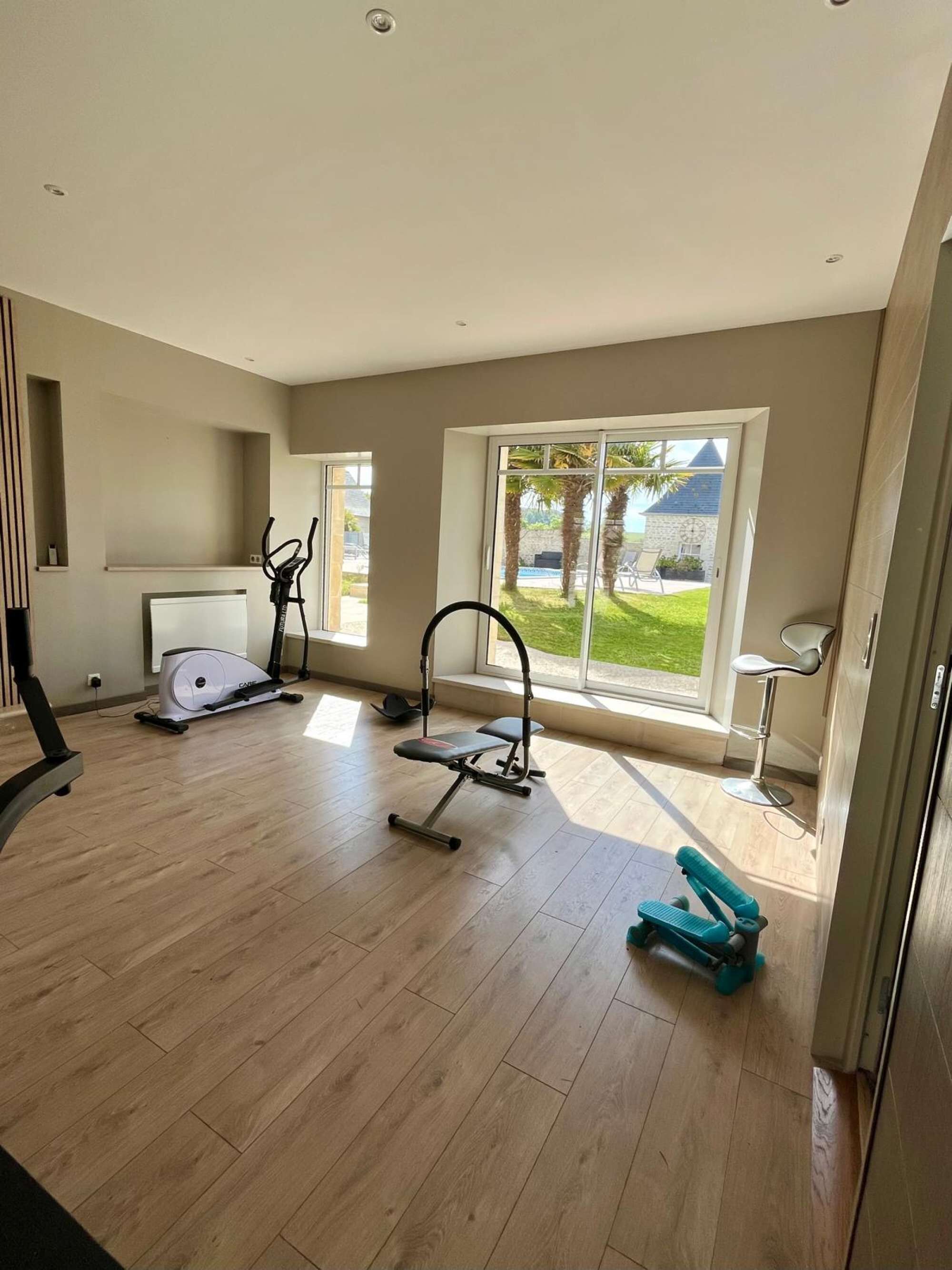 Une pièce spacieuse avec une grande fenêtre, un vélo d’appartement, un rameur et un petit banc d’exercices.