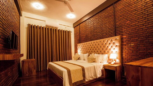 A bedroom with a wooden bed, a nightstand with a lamp, and a brick wall. - Manthra Leisure (Kandy)