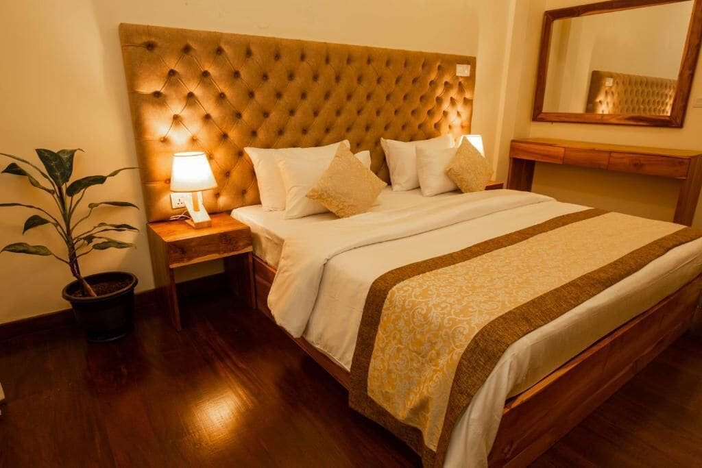 A hotel room with a tufted headboard, a bedside lamp, a potted plant, and a wooden nightstand.