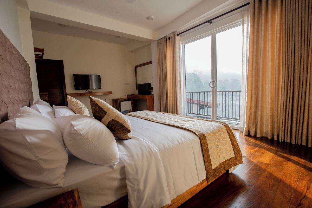A hotel room with a large bed, a television, a balcony with curtains, and a view of the outside.