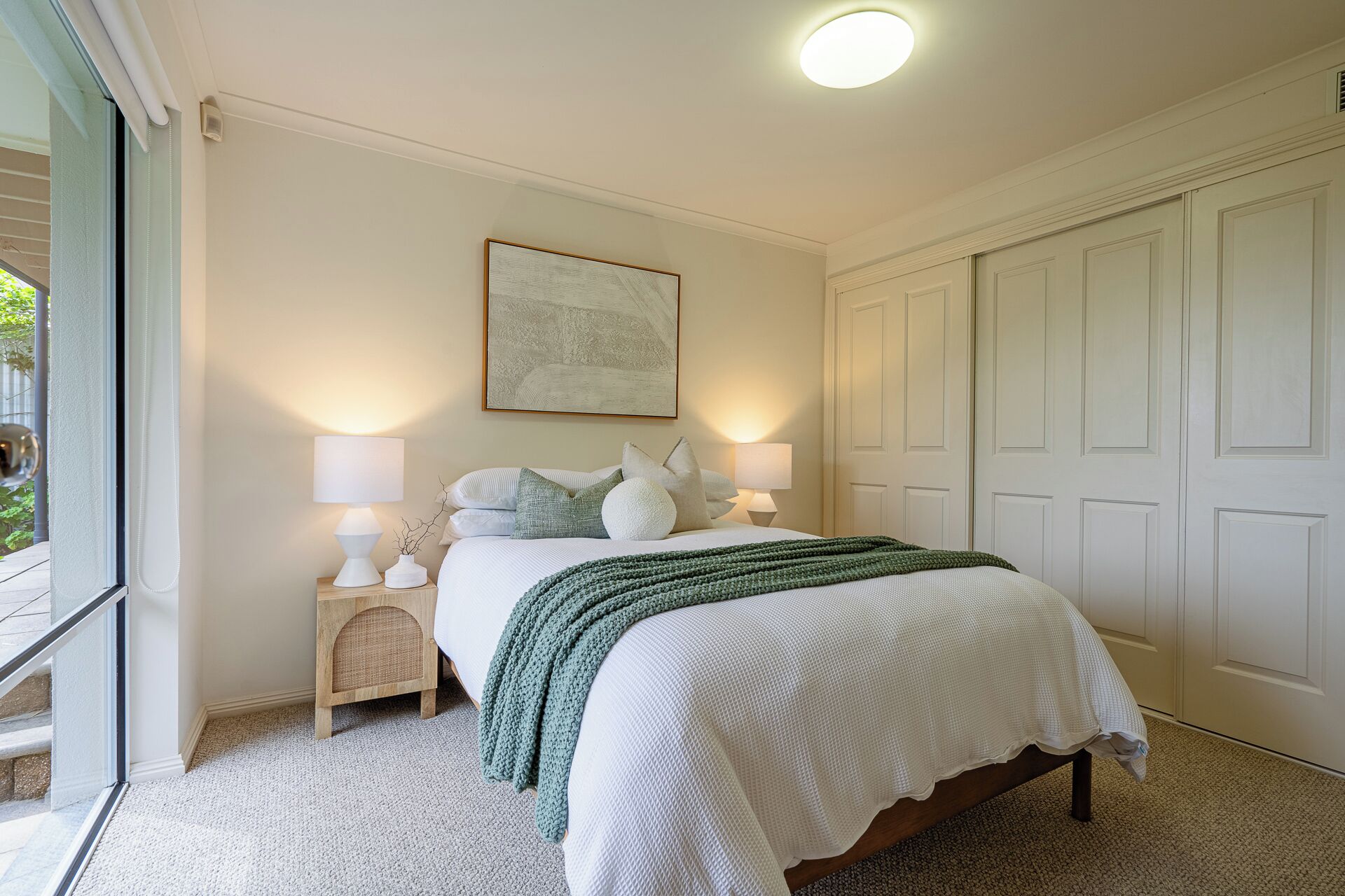 Une chambre à coucher avec un lit, deux tables de chevet avec des lampes, une grande fenêtre et une porte.