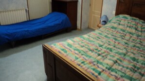 Une chambre avec deux lits, l’un recouvert d’une couverture bleue et l’autre d’une courtepointe multicolore. On y trouve une commode en bois et une lampe posée sur le sol.