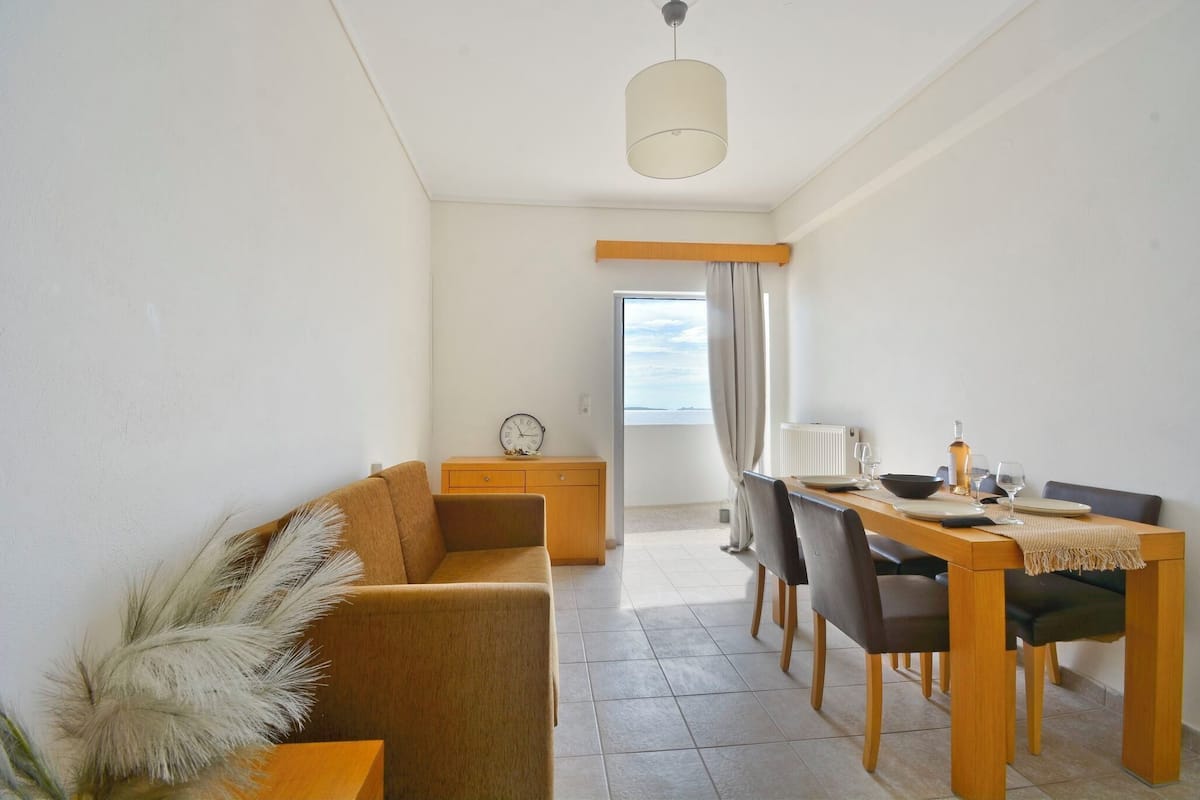 Uma sala de estar moderna com um sofá, mesa de jantar e cadeiras. Há uma janela com cortinas, um relógio em uma mesinha de apoio e um abajur sobre a mesa.