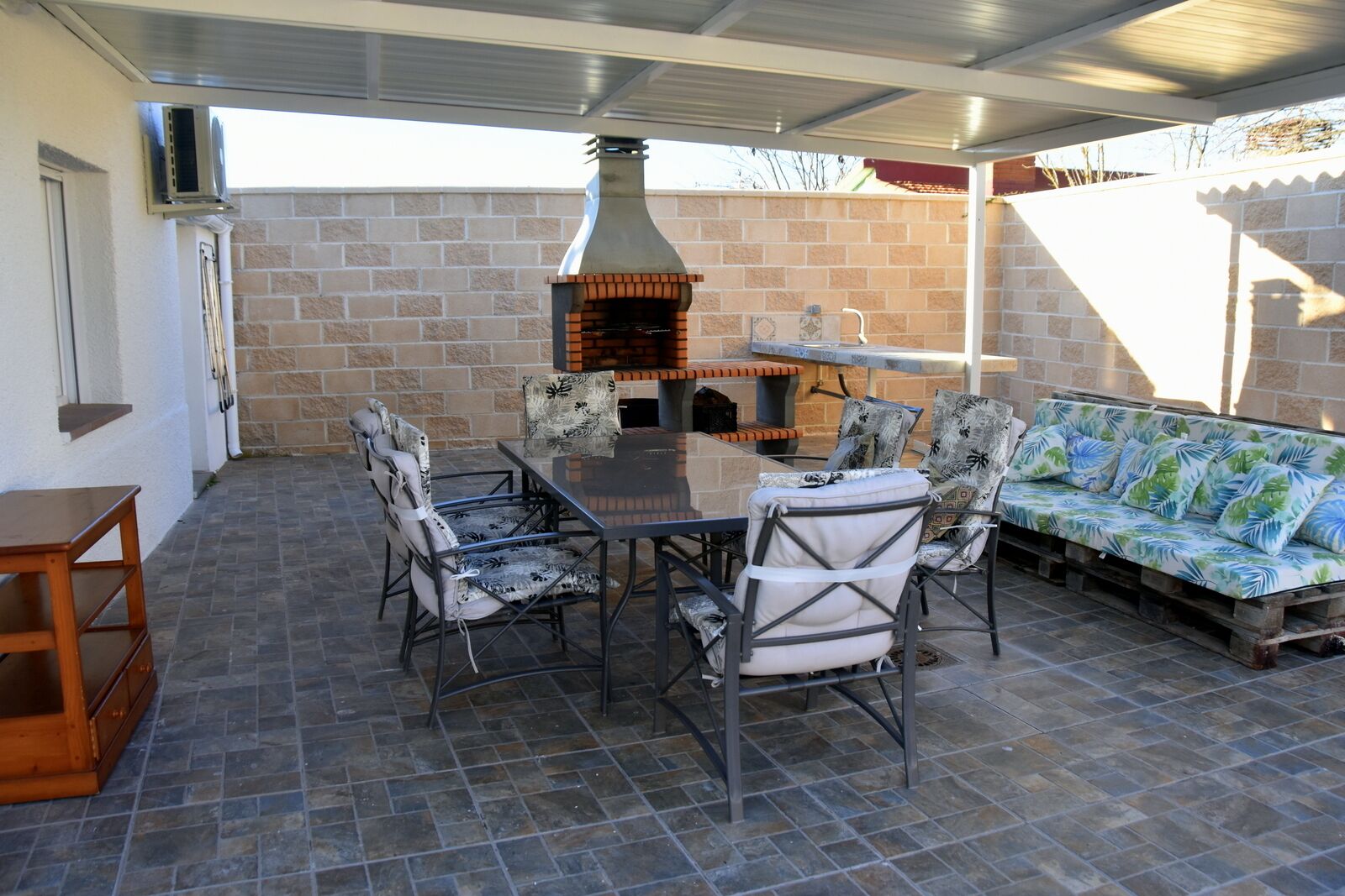 Eine Terrasse im Freien mit einer Backsteinmauer, einem Metallschutzdach, einem Backofen aus Backsteinen, einem Glastisch und einer Couch mit gemusterten Kissen.