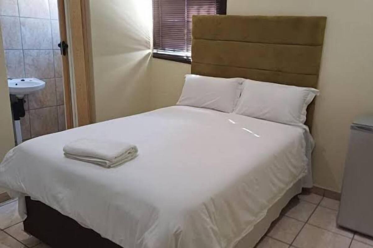 Een net opgemaakt bed met een opgevouwen witte handdoek, een klein nachtkastje en een compacte koelkast.