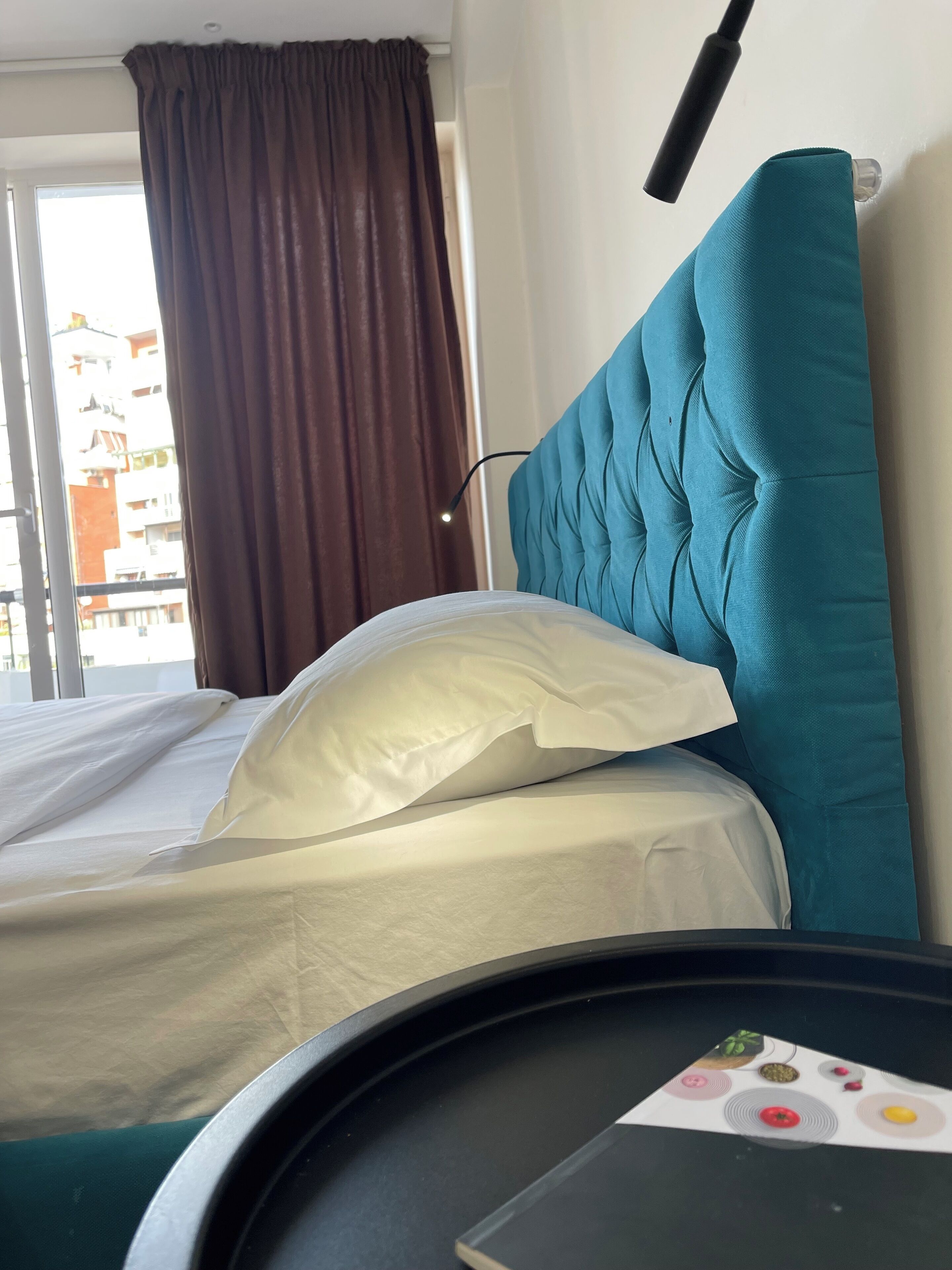 A hotel room with a bed, a headboard, a window with curtains, and a view of buildings outside.