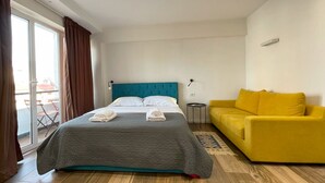 A hotel room with a bed, a yellow sofa, a small table, and a view of the outside. - Cozy studio with WiFi and AC in nice Tiranë (Tiranë)