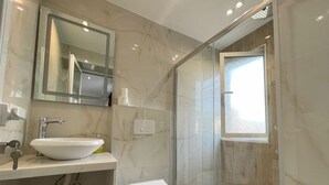 A modern bathroom with a white sink, mirror, and toilet. It features a glass shower enclosure and a window providing natural light. - Cozy studio with WiFi and AC in nice Tiranë (Tiranë)