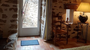 A rustic room with a stone wall, a wooden door leading outside, a chair, a table with a lamp, and a bed with a patterned cover. - LA TERRASSE DE PEYRE room "La Provençale (Comprégnac)