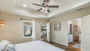 Un dormitorio con una cama grande, un ventilador de techo, una ventana con cortinas y un pasillo que conduce a un baño.