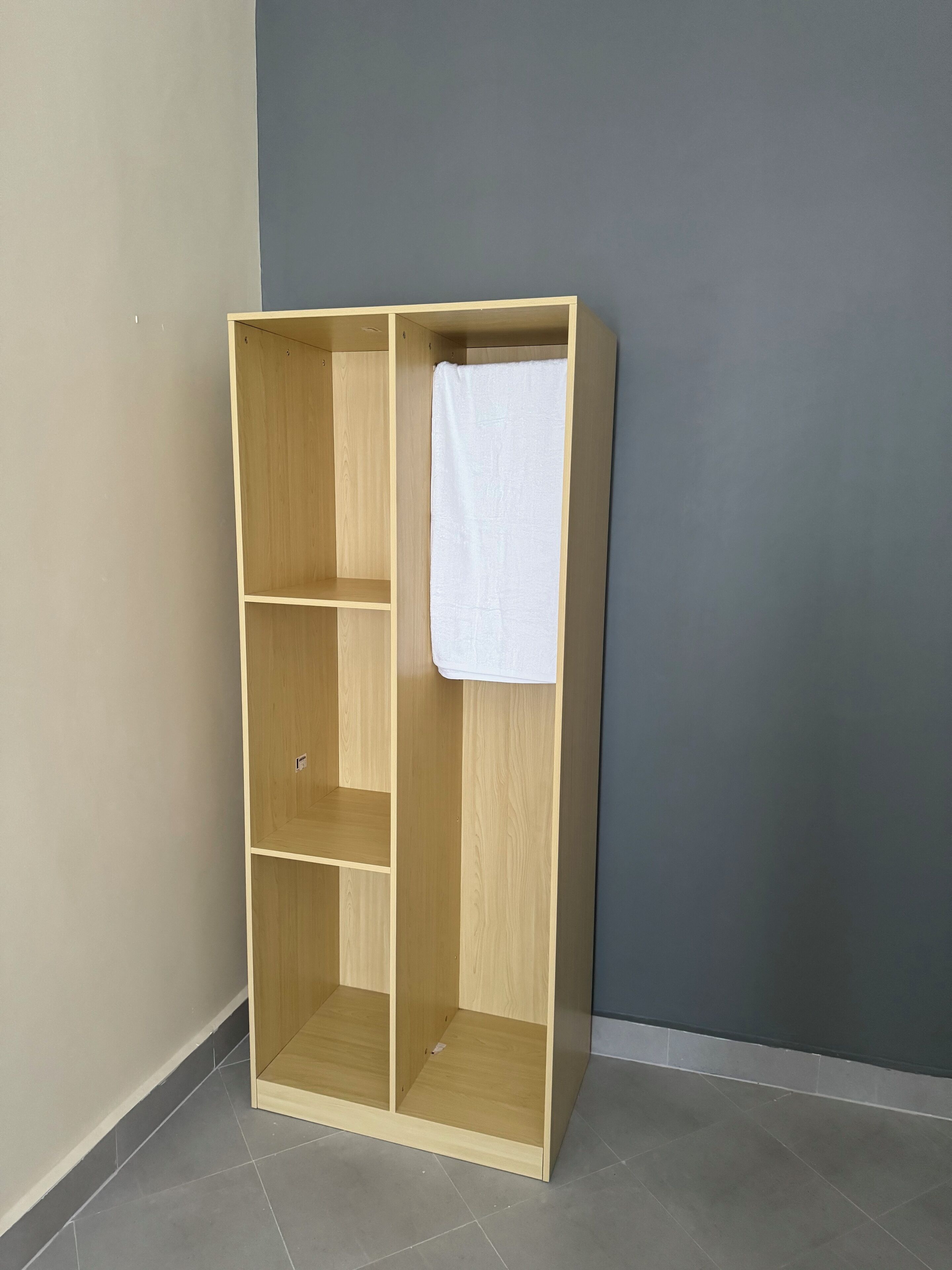 Ein Holzkleiderschrank mit einem weißen Handtuch, das an der Innentür hängt, steht in einer Ecke mit grauen Wänden und gefliestem Boden.
