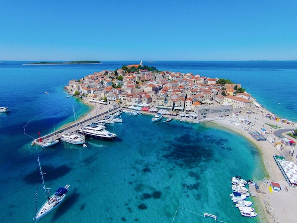 An aerial view of a coastal town with a marina, buildings with red-tiled roofs, and clear blue waters.