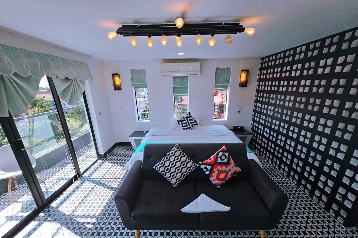 A room with a black sofa, patterned pillows, a bed with a geometric headboard, a balcony with patterned flooring, and a wall with a grid pattern.