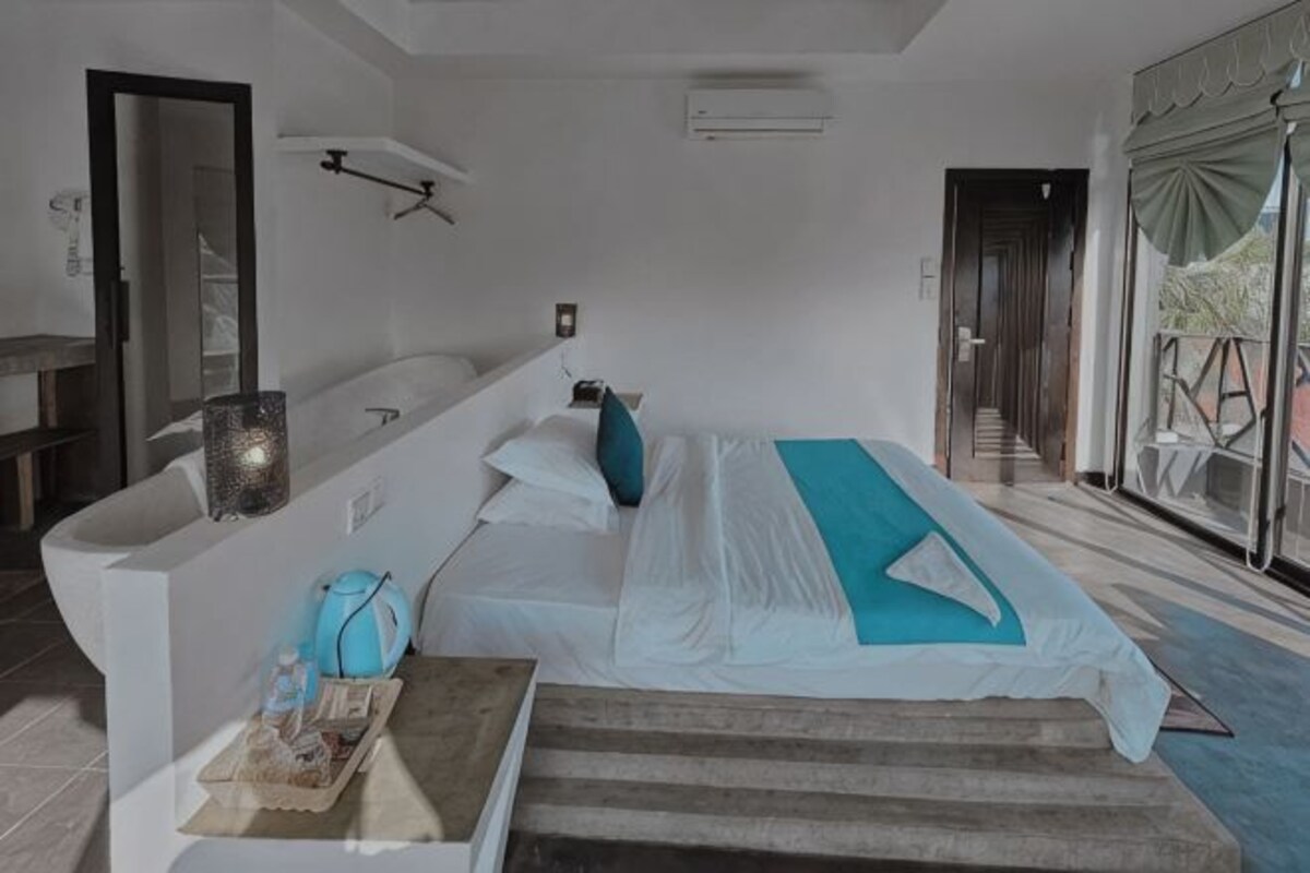 A modern hotel room with a bed, a sitting area, a bathroom, and a view of the outdoors.